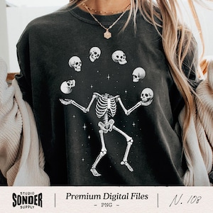 May include: Black graphic sweatshirt featuring a skeleton juggling skulls. The skeleton is white, with a detailed rib cage and limbs. The skulls are also white, with dark eye sockets. The design includes small white stars.