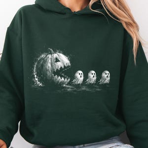 May include: A dark forest green hooded sweatshirt featuring a white graphic print of a large, jagged-mouthed jack-o'-lantern chasing three small, wide-eyed ghosts. The design has a textured, sketch-like appearance with splatters and shading.