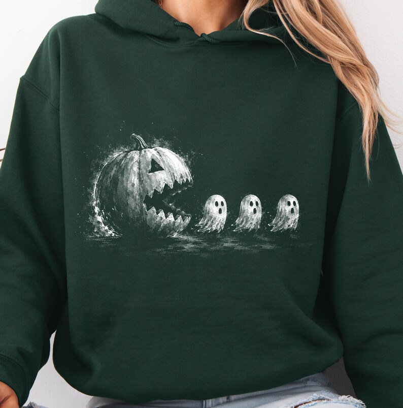 May include: A dark forest green hooded sweatshirt featuring a white graphic print of a large, jagged-mouthed jack-o'-lantern chasing three small, wide-eyed ghosts. The design has a textured, sketch-like appearance with splatters and shading.