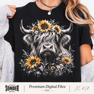 May include: A black t-shirt featuring a graphic illustration of a Highland cow with long horns. The cow is adorned with a crown of yellow sunflowers and white daisies, with additional sunflowers and wildflowers surrounding the lower portion.