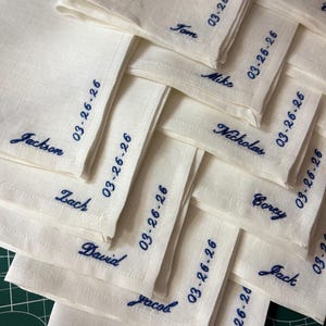 May include: A stack of off-white linen napkins embroidered with names and the date "03-26-26" in navy blue thread. Names include Tom, Mike, Nicholas, Corey, Jack, Jackson, Zach, David, Jacob, and Connor. The napkins are folded and stacked.