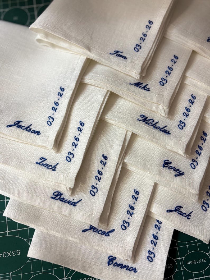 May include: A stack of off-white linen napkins embroidered with names and the date "03-26-26" in navy blue thread. Names include Tom, Mike, Nicholas, Corey, Jack, Jackson, Zach, David, Jacob, and Connor. The napkins are folded and stacked.