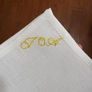 May include: A white linen napkin with gold embroidered initials "JOA" in a corner. The napkin has a simple, elegant design with a stitched border. The initials are in a cursive font.