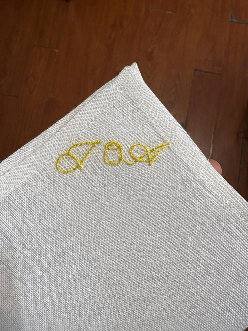 May include: A white linen napkin with gold embroidered initials "JOA" in a corner. The napkin has a simple, elegant design with a stitched border. The initials are in a cursive font.