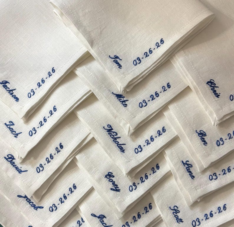 May include: A collection of folded white linen napkins, each embroidered with a name and the date 03-26-26 in navy blue thread. The names are in a cursive font and are different on each napkin. The napkins are neatly arranged.