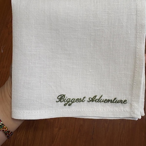 May include: A folded white linen handkerchief with the embroidered text "Biggest Adventure" in green script. The handkerchief is square and appears to be made of a textured fabric. The background is a wooden surface.