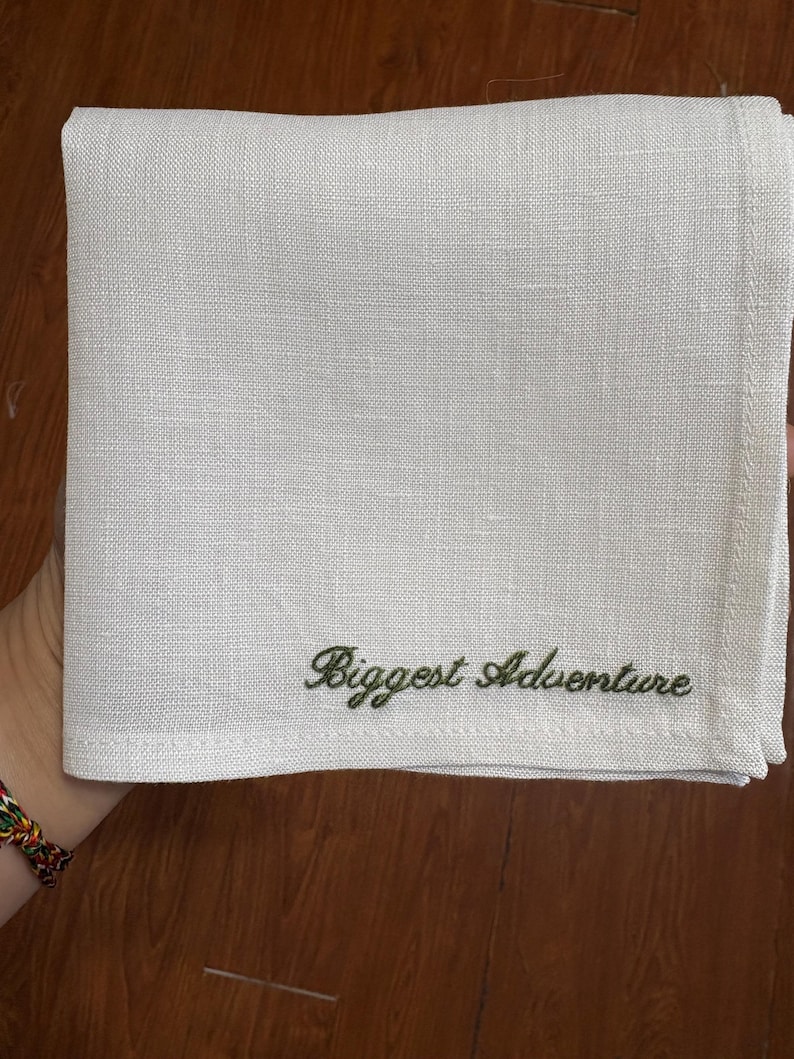 May include: A folded white linen handkerchief with the embroidered text "Biggest Adventure" in green script. The handkerchief is square and appears to be made of a textured fabric. The background is a wooden surface.