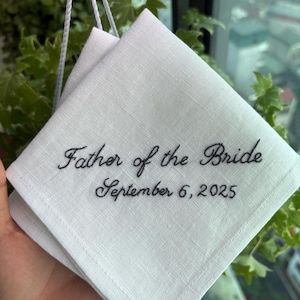 Könnte beinhalten: Weißes Leinen-Taschentuch mit der Aufschrift "Father of the Bride" und dem Datum "September 6, 2025" in schwarzer Schrift. Das Taschentuch ist gefaltet und vor einem Fenster mit Grün gehalten.