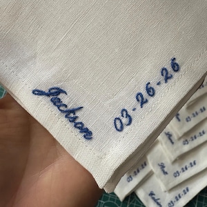 May include: White linen handkerchief with blue embroidered text. The handkerchief features the name "Jackson" and the date "03-26-26". Several other handkerchiefs with names and dates are visible in the background.