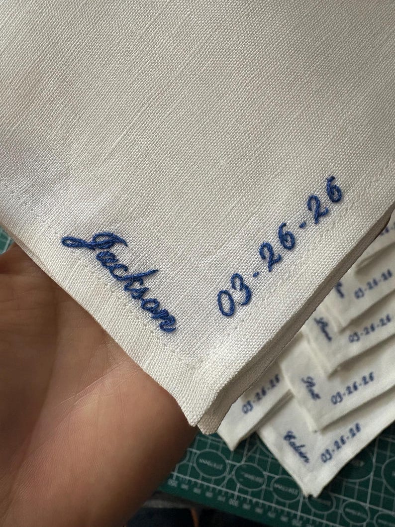 May include: White linen handkerchief with blue embroidered text. The handkerchief features the name "Jackson" and the date "03-26-26". Several other handkerchiefs with names and dates are visible in the background.
