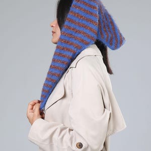 Puede incluir: Un gorro de punto a rayas azules y marrones con largas extensiones en forma de orejas. El gorro lo lleva una persona que lleva una gabardina beige. El diseño del gorro presenta rayas azules y marrones alternas.
