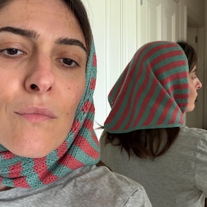Può includere: Un foulard in maglia a righe orizzontali in tonalità alternate di verde acqua spento e rosa corallo. Il tessuto presenta una lavorazione a maglia morbida e testurizzata che copre la parte superiore e i lati della testa, ricadendo sul collo.