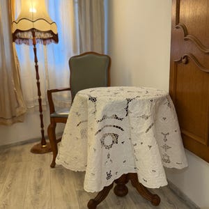 Op de afbeelding: Een ronde houten tafel met een centrale kolomvoet, bedekt met een wit rond kanten tafelkleed met gedetailleerde bloemmotieven en opengewerkt borduurwerk. De tafel heeft een centrale kolomvoet met gebogen poten in een kamer.