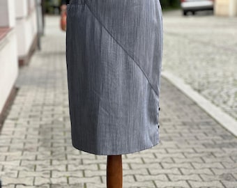 Op de afbeelding: Een grijze potloodrok op een houten mannequin. De rok heeft een diagonale naad over de voorkant en een kleine zak. Het heeft een getailleerde pasvorm en een knoopdetail aan de zijkant. De stof heeft een subtiel verticaal streepjespatroon.