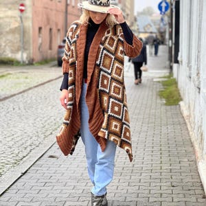 Può includere: Un lungo cardigan marrone e bianco a fantasia geometrica con un cappello abbinato. Il cardigan è aperto sul davanti e ha maniche lunghe. Il cappello è beige con una fascia decorativa. La persona indossa jeans blu e stivali argentati.
