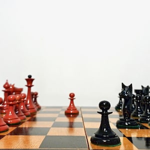 Puede incluir: Un juego de ajedrez con piezas rojas y negras dispuestas sobre un tablero de ajedrez de madera a cuadros. Las piezas rojas están a la izquierda, las piezas negras a la derecha y un peón rojo en el centro. Las piezas son brillantes y el tablero es marrón y negro.