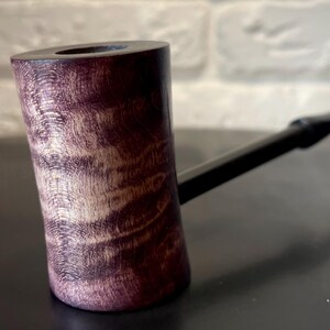 May include: A handcrafted tobacco pipe with a cylindrical bowl made of wood. The bowl has a dark purple and tan wood grain pattern. The stem is black and extends from the side of the bowl.