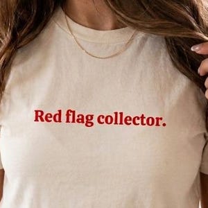 Könnte beinhalten: Ein kurzärmeliges, cremeweißes T-Shirt mit Rundhalsausschnitt und dem Schriftzug "Red flag collector." in einer kräftigen, roten Serifenschrift auf der Brust. Das Kleidungsstück hat eine klassische Passform und ein schlichtes Design.