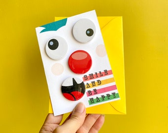 Birthday badge greetings card. Smile & be happy. Pin button badge gift. Clown, funny face theme, googley eyes, red nose