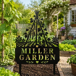 May include: A black metal garden sign featuring a cutout design of a tripod trellis with climbing bean plants and the text MILLER GARDEN. The sign includes two small heart-shaped cutouts and is supported by two stakes inserted into the soil.