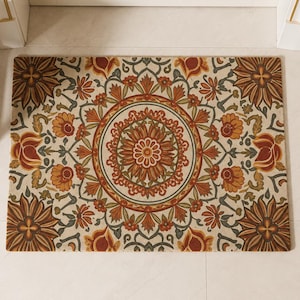 Puede incluir: Un felpudo rectangular con un patrón de mandala circular central rodeado de motivos florales. El diseño incorpora tonos de naranja quemado, rojo intenso, verde apagado y crema, con grandes formas de flores situadas en cada esquina.