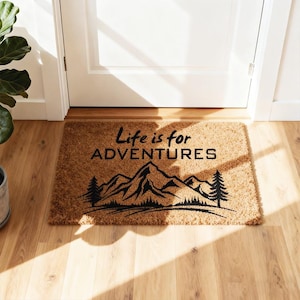 May include: A rectangular doormat made of natural fibres. The mat features a black graphic of mountains and trees, with the phrase "Life is for ADVENTURES" in black text. The mat is placed in front of a white door.
