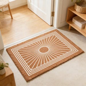 Op de afbeelding: Bruine rechthoekige deurmat met een wit sunburst-ontwerp. De mat heeft een centrale cirkel met stralende lijnen, omlijst door een witte rechthoekige rand. De mat ligt voor een witte deur.