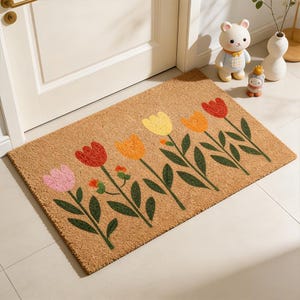 Può includere: Un zerbino rettangolare in fibra naturale, con una fila di tulipani colorati in rosa, rosso, giallo e arancione. I tulipani hanno steli e foglie verdi. Il tappeto è posto davanti a una porta bianca.