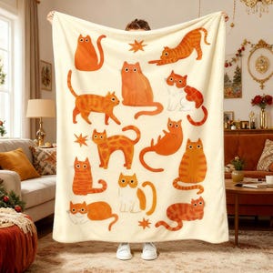 May include: A cream-coloured blanket adorned with orange cat illustrations in diverse poses. The cats display varied patterns and expressions, some with star motifs. The blanket is held up, highlighting its design and soft texture.