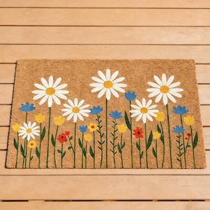 Op de afbeelding: Een rechthoekige deurmat van natuurlijke kokosvezel. De mat heeft een bloemenontwerp met grote witte madeliefjes, gele en blauwe wilde bloemen en groene stelen tegen een bruine achtergrond. De mat ligt op een houten oppervlak.