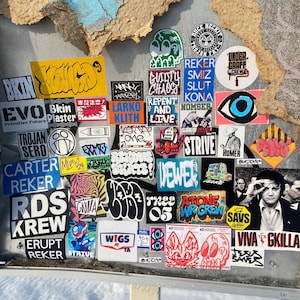 May include: A rectangular display board covered in a dense collage of various colorful stickers featuring diverse typography, graphic illustrations, and text including BKIN, GUILTY CHAOS, REKER, STRIVE, RDS KREW, and a black and white portrait of Elvis.
