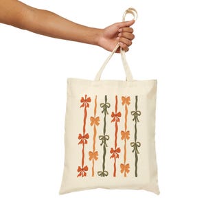 May include: A cream-colored tote bag with a repeating pattern of vertical lines adorned with bows in shades of orange, red, and green. The bag is held by a person's hand, showcasing its design and carrying capacity.
