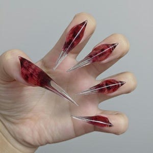 Może przedstawiać: Dłoń prezentująca długie, spiczaste paznokcie akrylowe. Paznokcie są w głębokim czerwonym kolorze z przezroczystą, ostrą końcówką. Projekt daje wygląd estetyki przypominającej pazur.