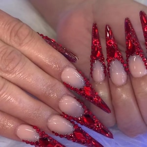 May include: Long, pointed red acrylic nails with a clear base and red gem accents. The nails are decorated with various sizes of red gems, creating a glamorous design. The nails are on a white surface.