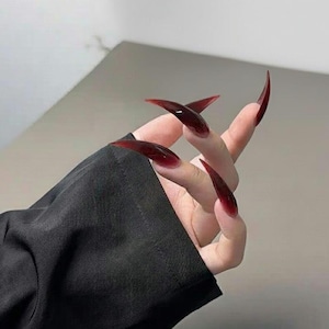 Puede incluir: Una mano con uñas acrílicas largas y puntiagudas de color burdeos. Las uñas son de un rojo intenso y brillante, y la mano está parcialmente cubierta por una manga negra. Las uñas tienen una forma afilada y curvada.