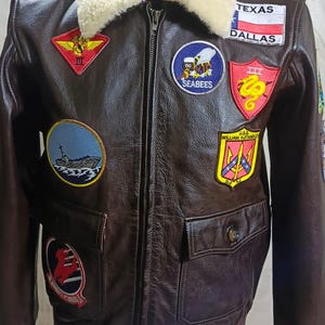 Könnte beinhalten: Braune Lederjacke mit cremefarbenem Lammfellkragen. Die Jacke hat mehrere Aufnäher, darunter eine Texas-Flagge und ein "SEABEES"-Aufnäher. Sie hat einen Reißverschluss und zwei Vordertaschen.