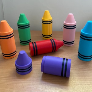 May include: A collection of colorful crayon-shaped containers in orange, green, yellow, pink, blue, red, and purple. Each container has black bands around the body. The containers are arranged on a light brown surface.