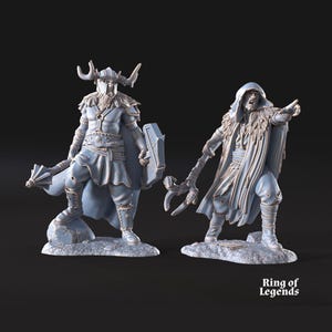 May include: Two light grey fantasy miniature figurines standing on rocky bases. The left figure wears a horned helmet, cape, and holds a mace and shield. The right figure wears a hooded cloak, fur collar, and holds a staff. Text reads Ring of Legends.