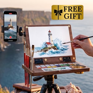 Könnte beinhalten: Eine tragbare Kunststaffelei aus Holz auf einem Stativ mit integrierter Aquarellpalette, Pinselhaltern und einer Smartphone-Halterung. Die Staffelei zeigt ein Leuchtturm-Gemälde, einen Pinsel in Gebrauch und eine Muschel auf der Ablage.