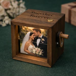以下が含まれることがあります： 結婚式の写真が入った木製のフォトキューブ。キューブの上部には「Together forever」の文字が刻まれています。上部には小さな金色のボタンがあり、側面には木のクランクがあります。写真は新郎新婦です。