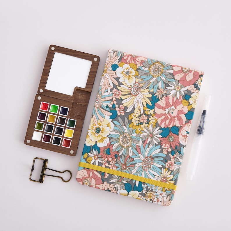 May include: A wooden watercolor palette with a selection of vibrant paints, a floral patterned notebook with a yellow elastic band, a water brush, and a binder clip. The palette is open, revealing a white mixing area.