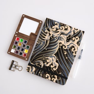 May include: A wooden watercolor palette with 16 colored paint squares, a notebook with a wave pattern, a water brush, and a binder clip. The notebook has a black elastic band. The palette has a built-in mixing area.