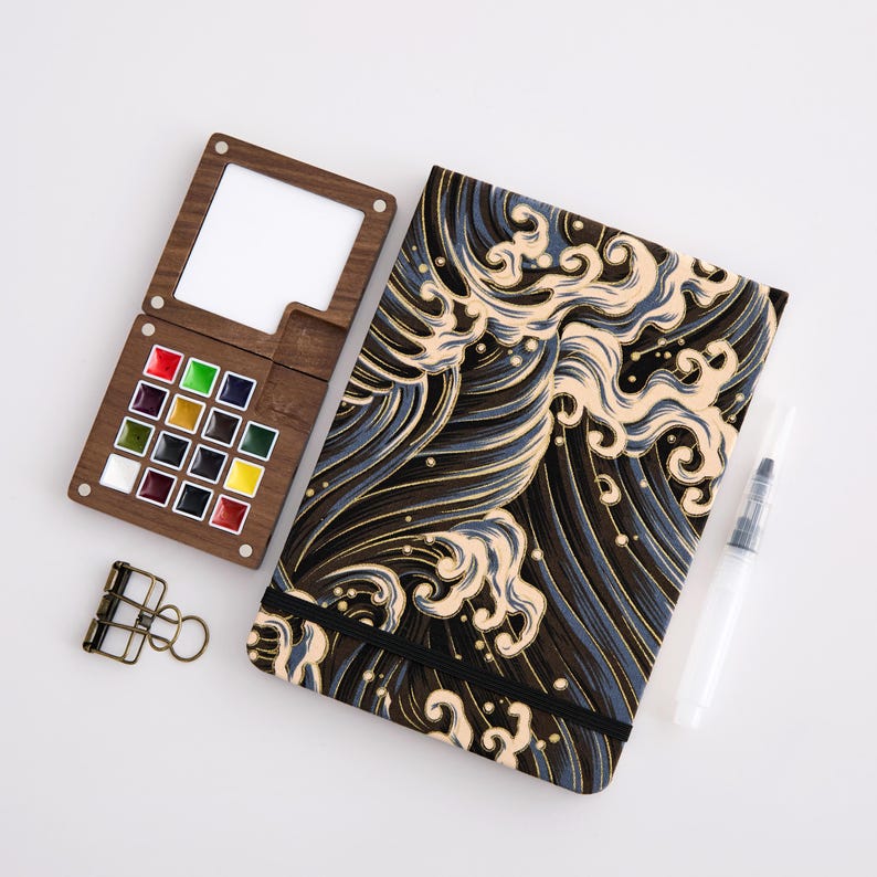 May include: A wooden watercolor palette with 16 colored paint squares, a notebook with a wave pattern, a water brush, and a binder clip. The notebook has a black elastic band. The palette has a built-in mixing area.