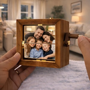 May include: A wooden photo cube with a family portrait visible through a window. The cube has a crank on the side and is held by a person's hand. The photo shows a smiling family of five. The cube is made of dark wood.