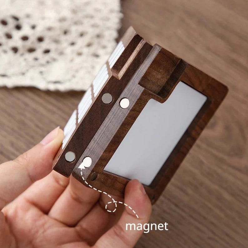 May include: A dark brown wooden card holder with a white interior and a series of magnets. The holder is rectangular with a layered design. The word "magnet" is written in white.