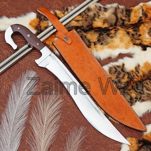 May include: A curved blade knife with a dark wood handle and a polished steel blade. The knife is accompanied by a brown leather sheath. The text "Zaime W" is visible on the blade. The knife is displayed on a brown surface.