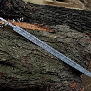 May include: A steel sword blade with a patterned design rests on a textured, dark brown log. The blade has a pointed tip and a long, narrow profile. The text "Zamie Wzd" is visible on the blade.