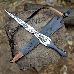 May include: A silver-toned metal dagger with a pointed blade and a black handle featuring decorative cutouts, accompanied by a black leather sheath embossed with a rose design and accented with a brown leather tip and a black strap.