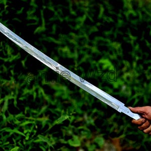 Könnte beinhalten: Eine lange, silberne Katana-Klinge mit rechteckigem Griff. Die Klinge wird von einer Hand vor einem Hintergrund aus grünem Laub gehalten. Die Klinge ist etwa 76 cm lang.