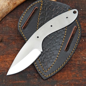 May include: A silver-colored metal knife blade blank with a curved handle section featuring three small circular holes and a pointed tip, resting on a black leather sheath with a textured pattern and yellow stitching along the edges.